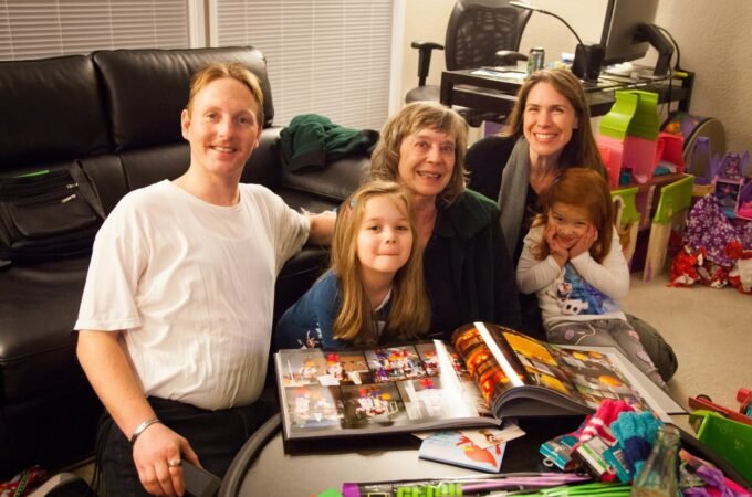 A group gathers in a living room to review photo albums. Someone holds a little one while colorful gifts sit nearby.