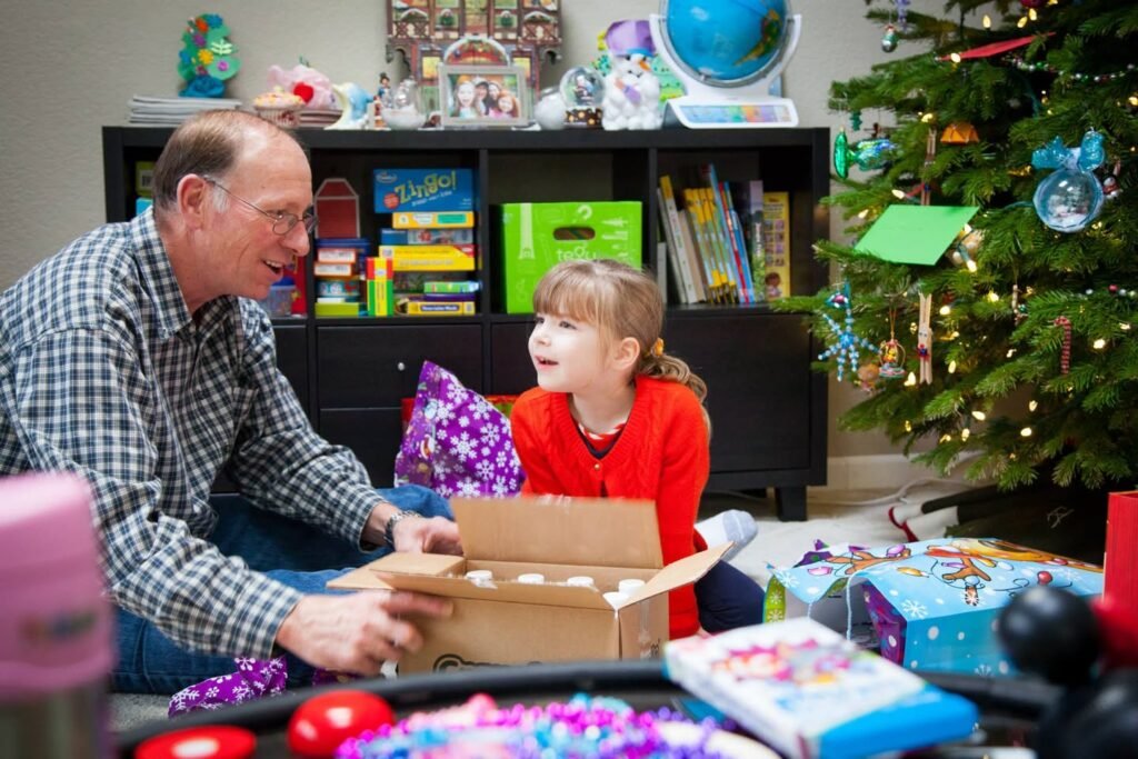 A child excitedly opening a gift box with an adult near a decorated Christmas tree.