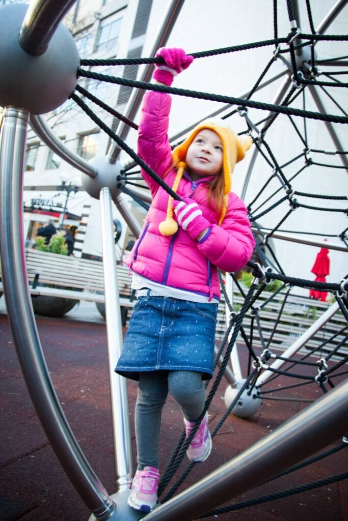 A child in a bright pink jacket and orange beanie holds onto a geodesic dome climbing structure.