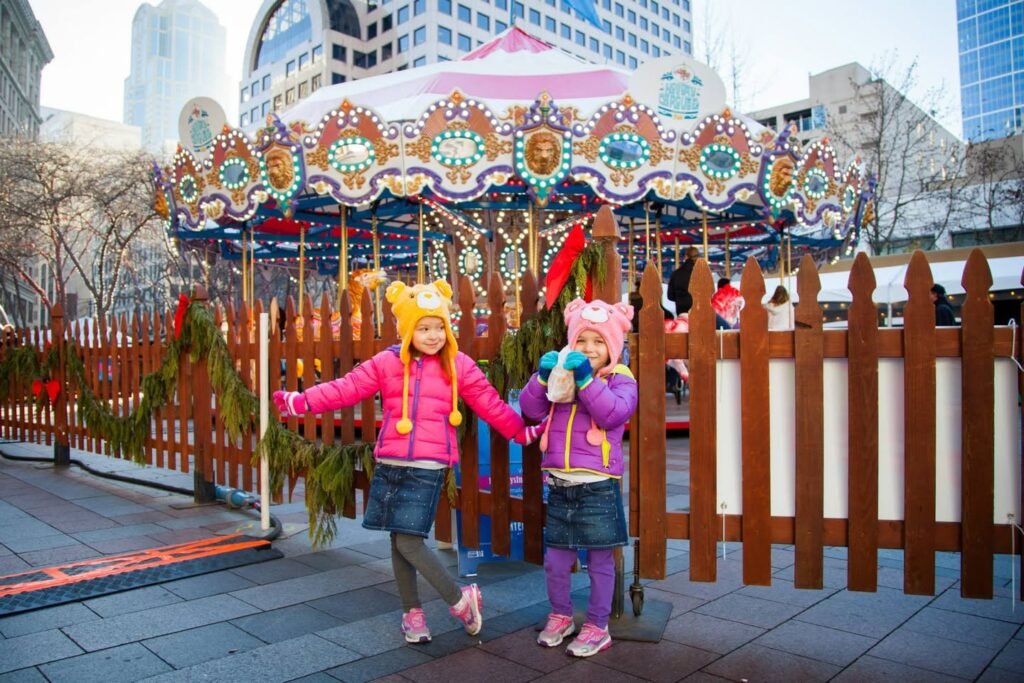 Two children in colorful winter hats stand in front of an ornate carousel surrounded by festive decorations.