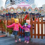 Two children in colorful winter hats stand in front of an ornate carousel surrounded by festive decorations.