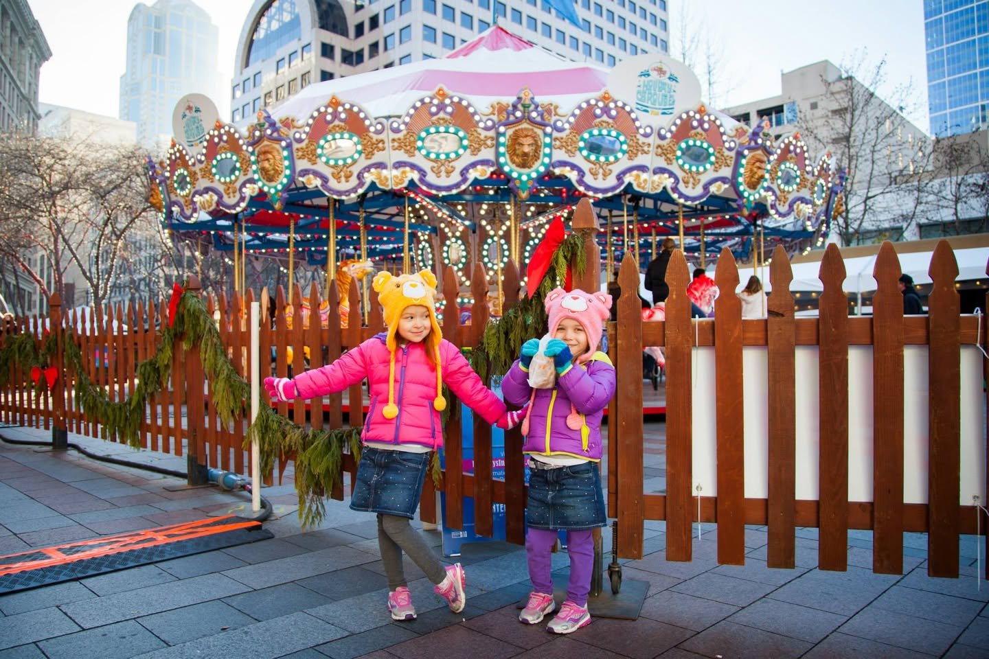 New Year’s Day in Seattle Two children in colorful winter hats stand in front of an ornate carousel surrounded by festive decorations.