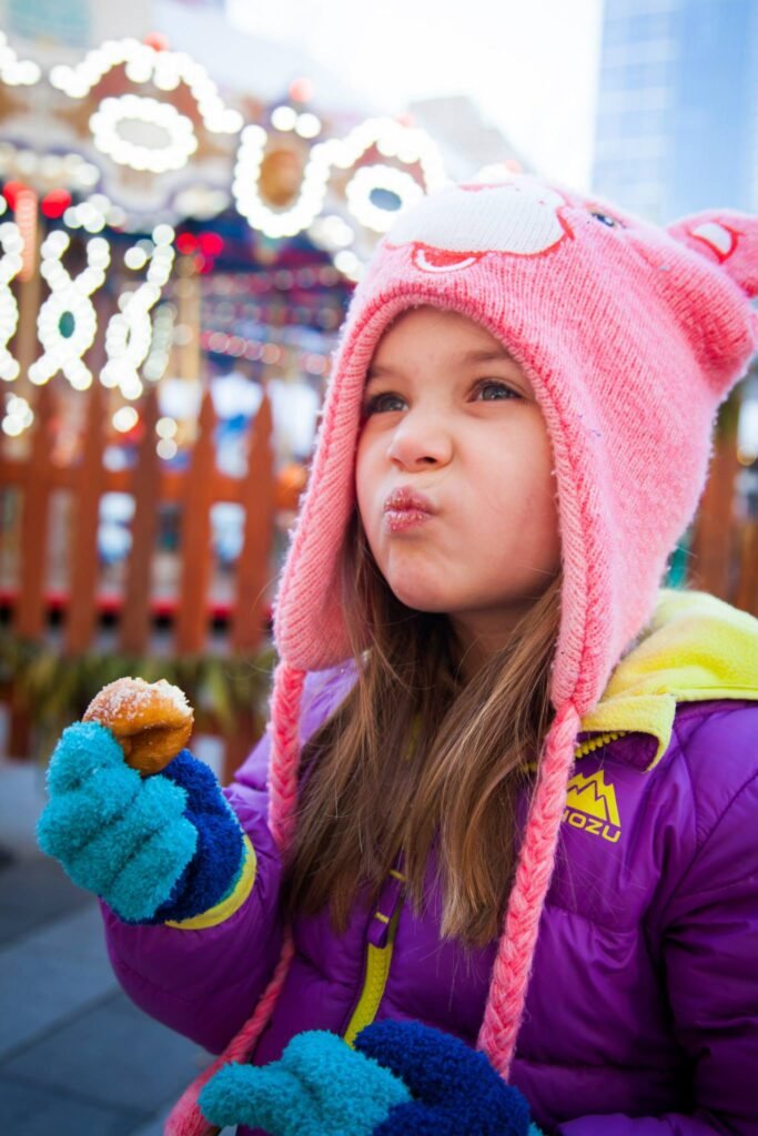 A close-up of a person in a pink hat holding a pastry with blurred Wintermarket lights in the background.
