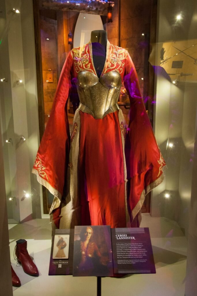 A close-up view of a dramatic red and gold period costume displayed in a museum exhibit case, accompanied by informational placards