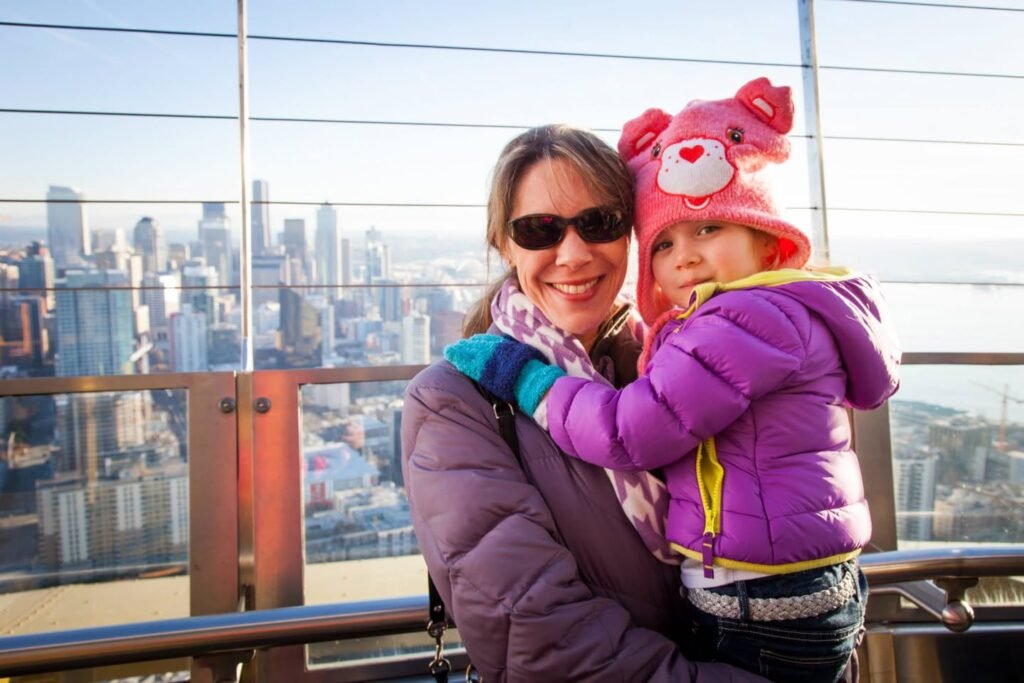 A warm embrace at the Edge with A Blue Star, capturing the stunning Seattle skyline with panoramic views. Both wearing warm jackets against a clear blue sky.
