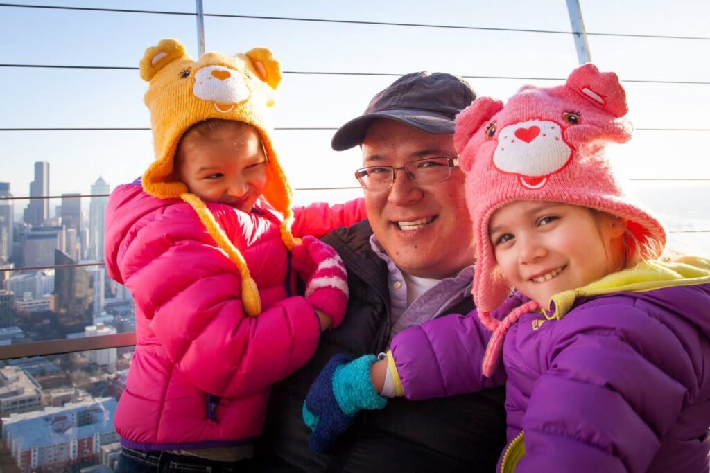 A man holding two young children wearing colorful winter jackets and animal-themed hats at an outdoor observation deck. The children's bright clothing contrasts with the cityscape in the background.
