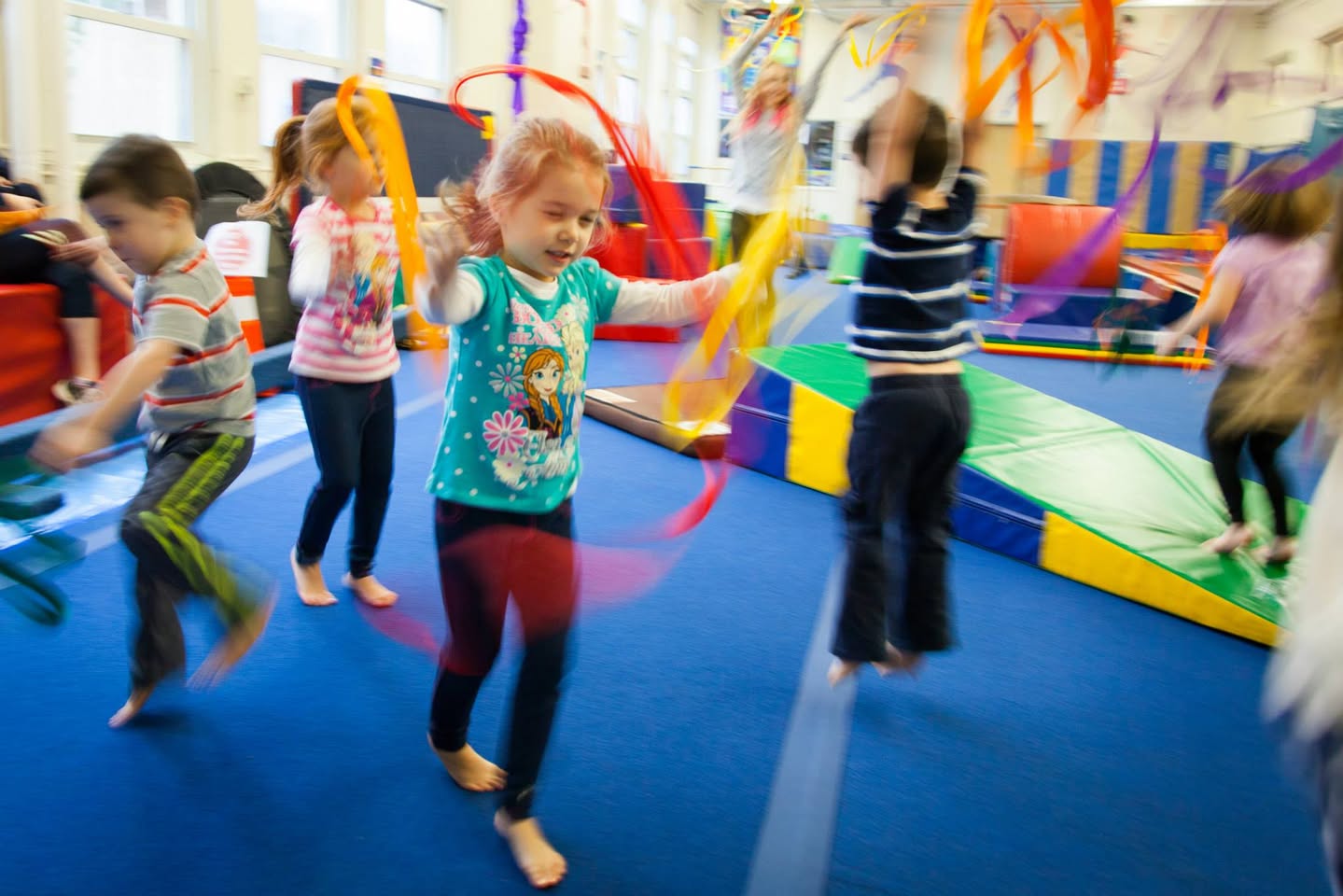 Children jump on a blue mat while playing with vibrant streamers in an indoor gym.