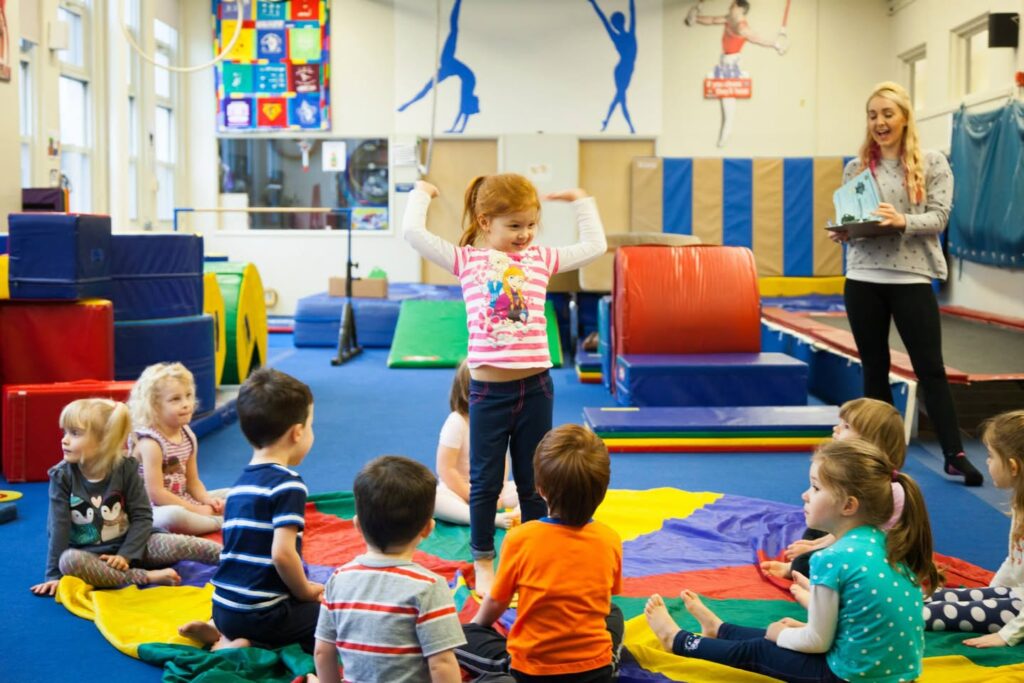 A young child performs a tumble in a colorful indoor gym while an instructor and other children watch from the mats.