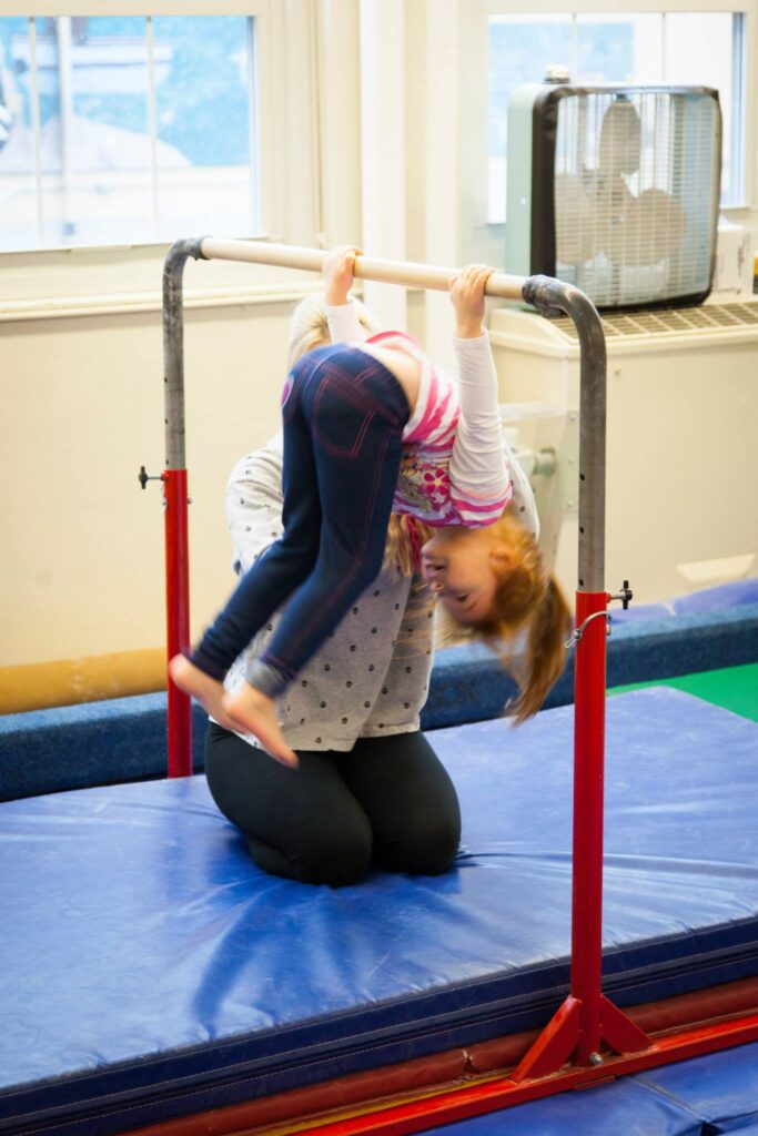 A child performs an inverted gymnastics move on bars in a gym with blue mats and snow visible outside the windows.