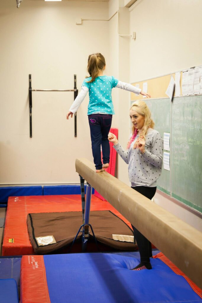 A person balances on a low beam with a coach assisting during a gymnastics lesson in a brightly lit gym.