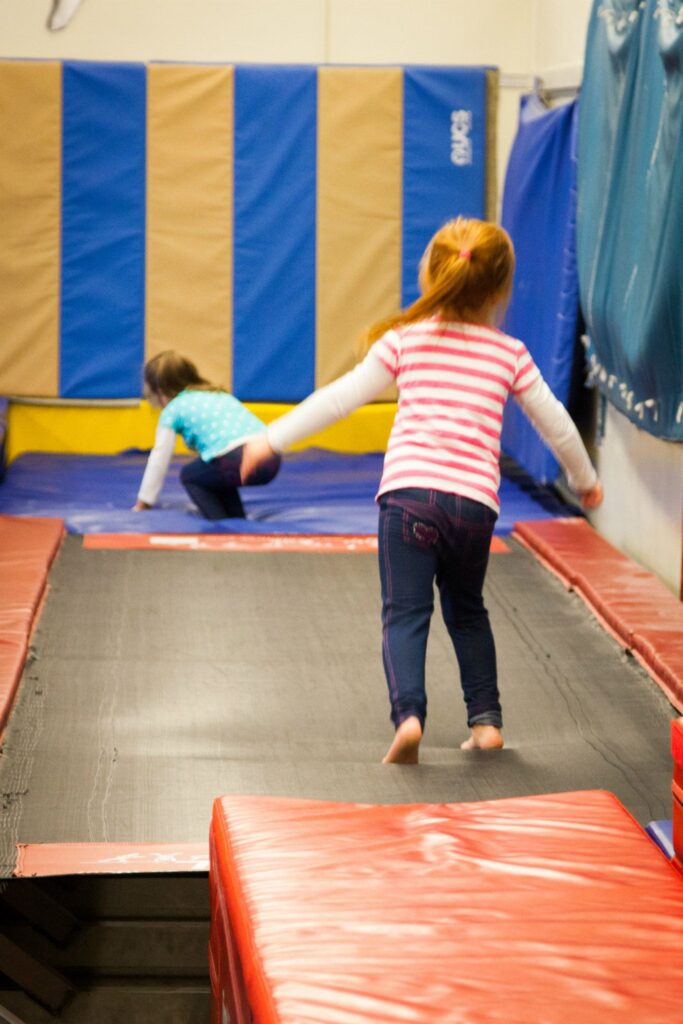 Two children bounced and played on an indoor trampoline with a blue and yellow padded wall.