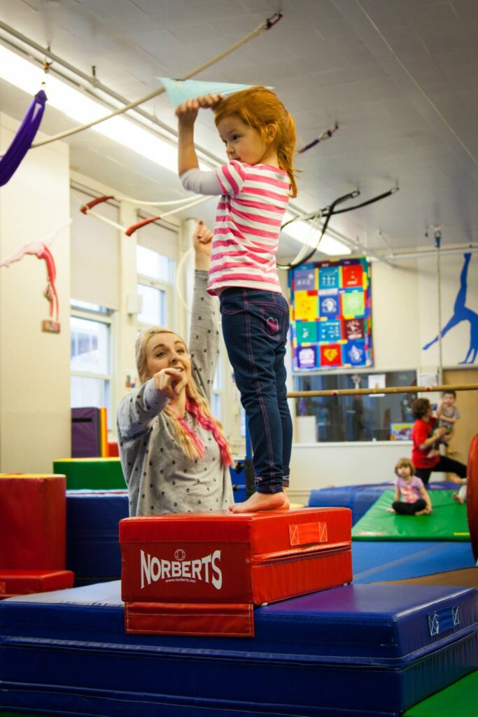 A young child stands on a red 'Norberts Gymnastics' balance box, supported by an adult, inside a bright, colorful gymnastics training studio.