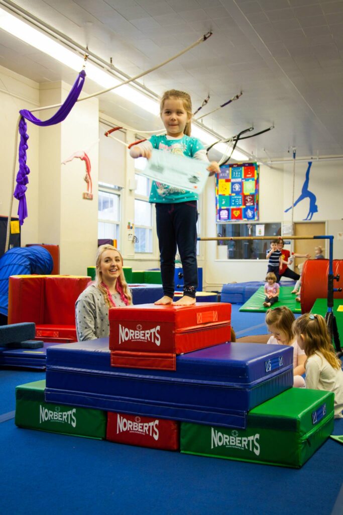 A child balances on a colorful platform in a children's gym, with a purple silk in the background.