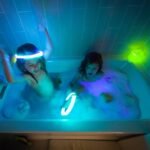 Children playing with bright glow sticks and toys in a dark bathroom tub.