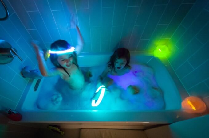 Children playing with bright glow sticks and toys in a dark bathroom tub.