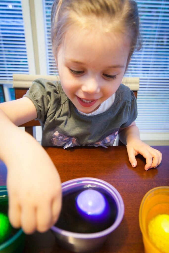 A child smiles while mixing colorful liquids in bowls on a kitchen table.