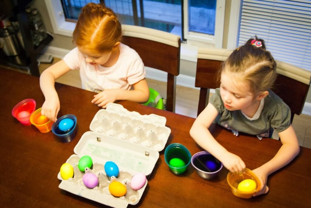 Two children dipping Easter eggs into colorful dye cups at a kitchen table.