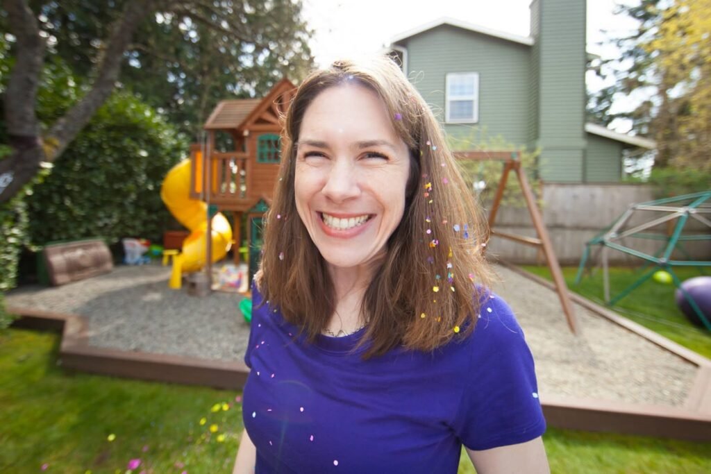 A smiling woman in a blue shirt stands in a backyard filled with play equipment and greenery amid floating confetti.