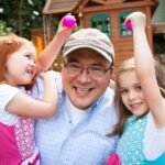 A smiling adult and two children playing outdoors in a backyard, each holding a pink ball with a wooden playhouse in the background.