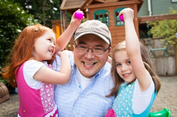 A smiling adult and two children playing outdoors in a backyard, each holding a pink ball with a wooden playhouse in the background.