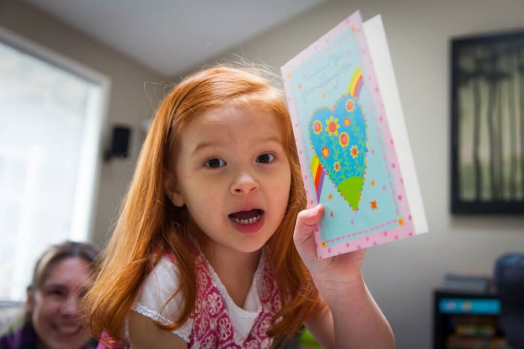 A person with red hair holds up a colorful handmade card featuring a blue heart design. Another individual is visible smiling in the background of a home interior.