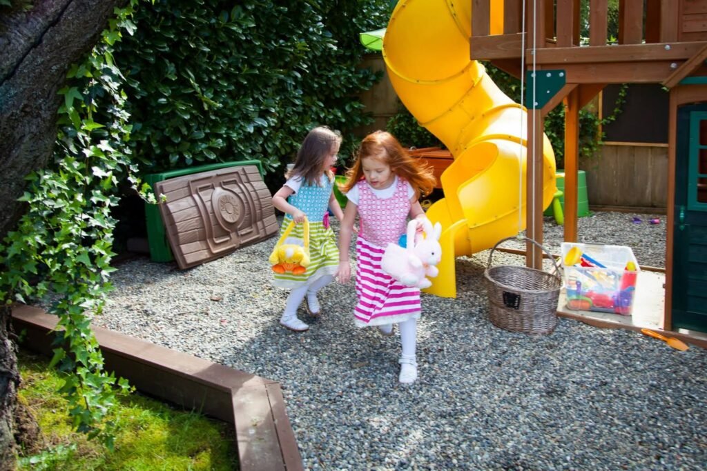 Two children walk on a gravel surface beside a yellow slide and lush greenery.