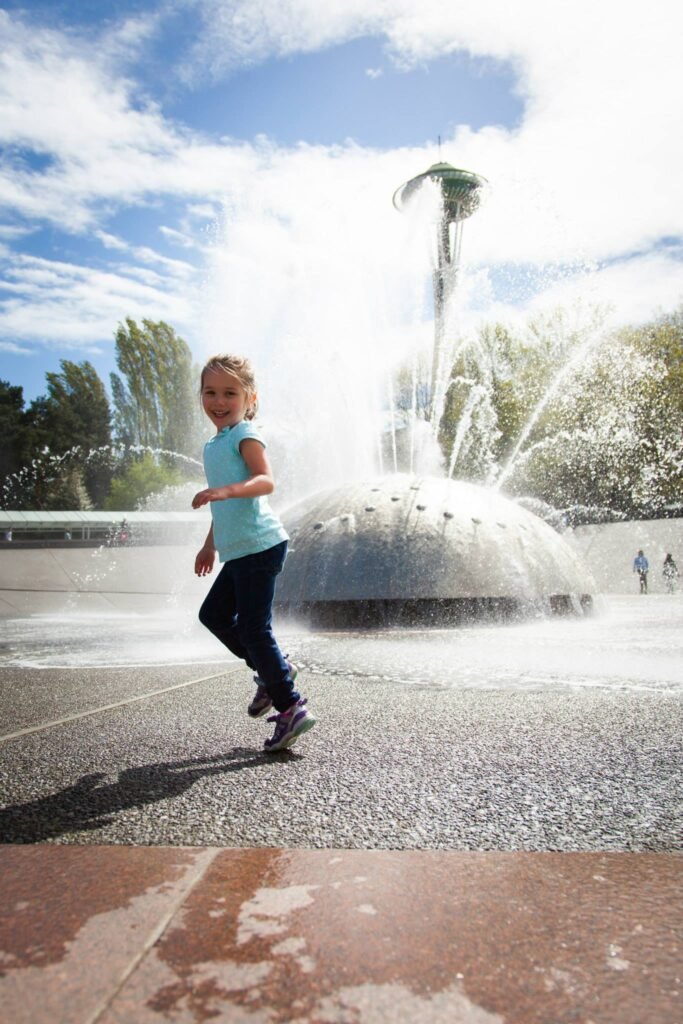 A young child plays near a large fountain in Seattle, with the Space Needle visible against a bright, partly cloudy sky.
