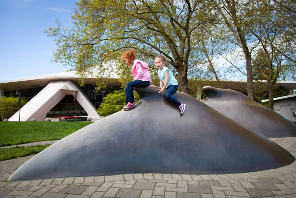 Children exploring a large metal sculpture at the Amos Rex contemporary art museum. Palm Hall architecture and trees in the background.