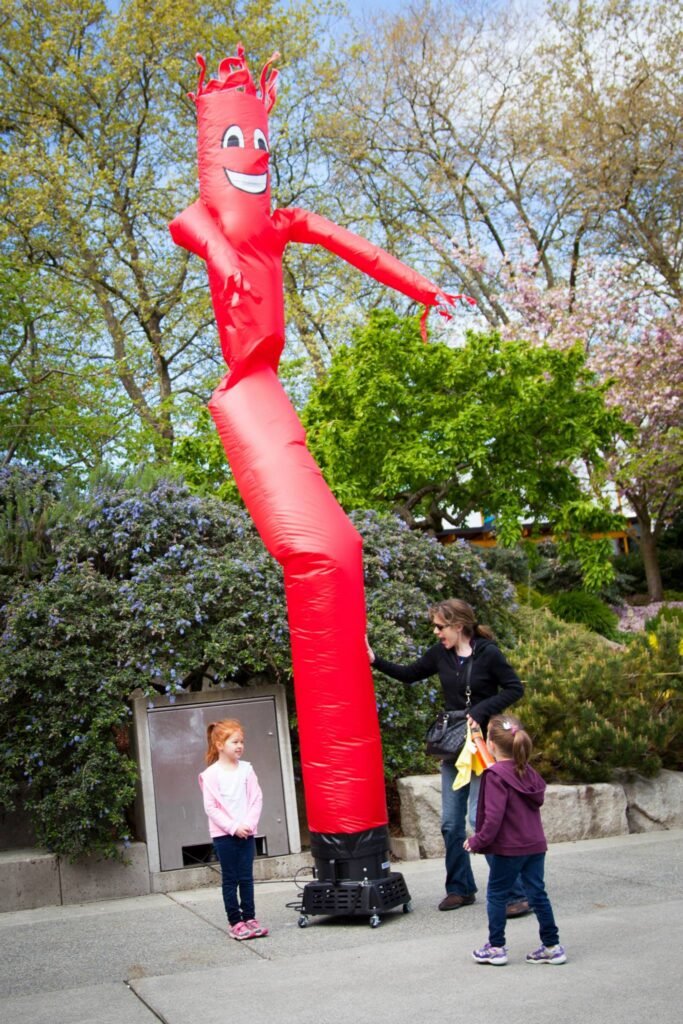 A smiling giant red inflatable character towers over a family at a public park during the spring. Blossoming cherry trees provide a picturesque background to the day's fun activities.