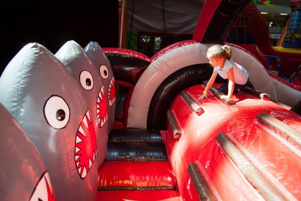 A child plays on a red and white inflatable with large gray shark decorations. The bouncy structure features cartoonish shark mouths and fins.