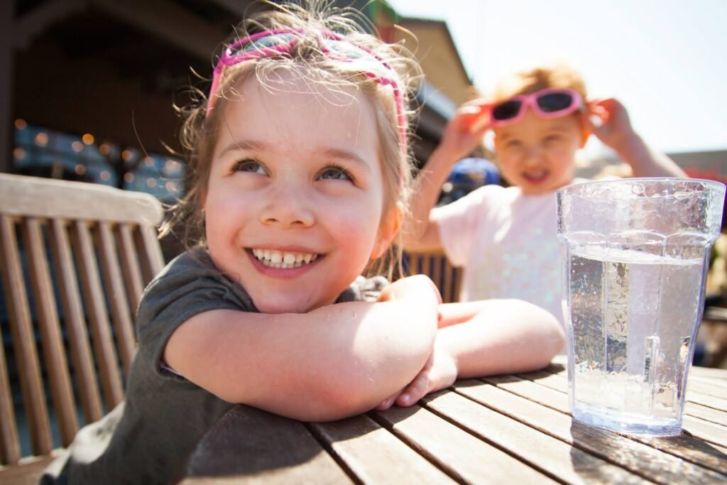 A child leans on a wooden table, smiling at the camera while another adjusts sunglasses.