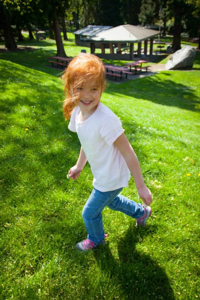A child in a white shirt runs across a grassy park area, with a scenic gazebo and trees visible in the background.