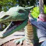 A child wears pink sunglasses and hugs a large green dragon statue in a sunny park setting.