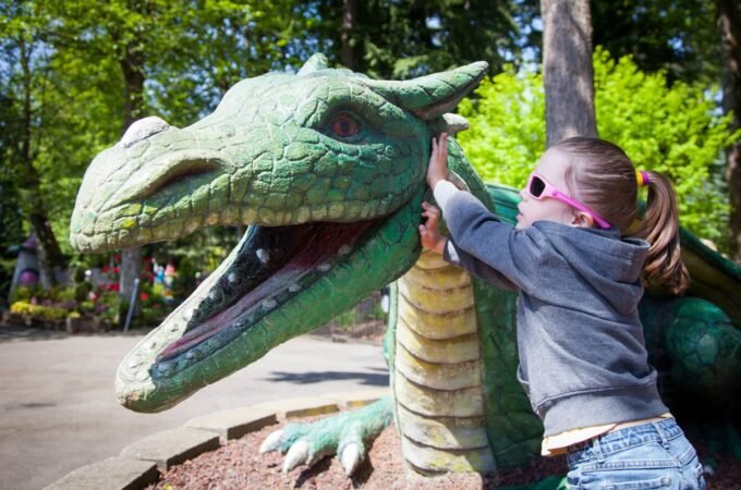 A child wears pink sunglasses and hugs a large green dragon statue in a sunny park setting.