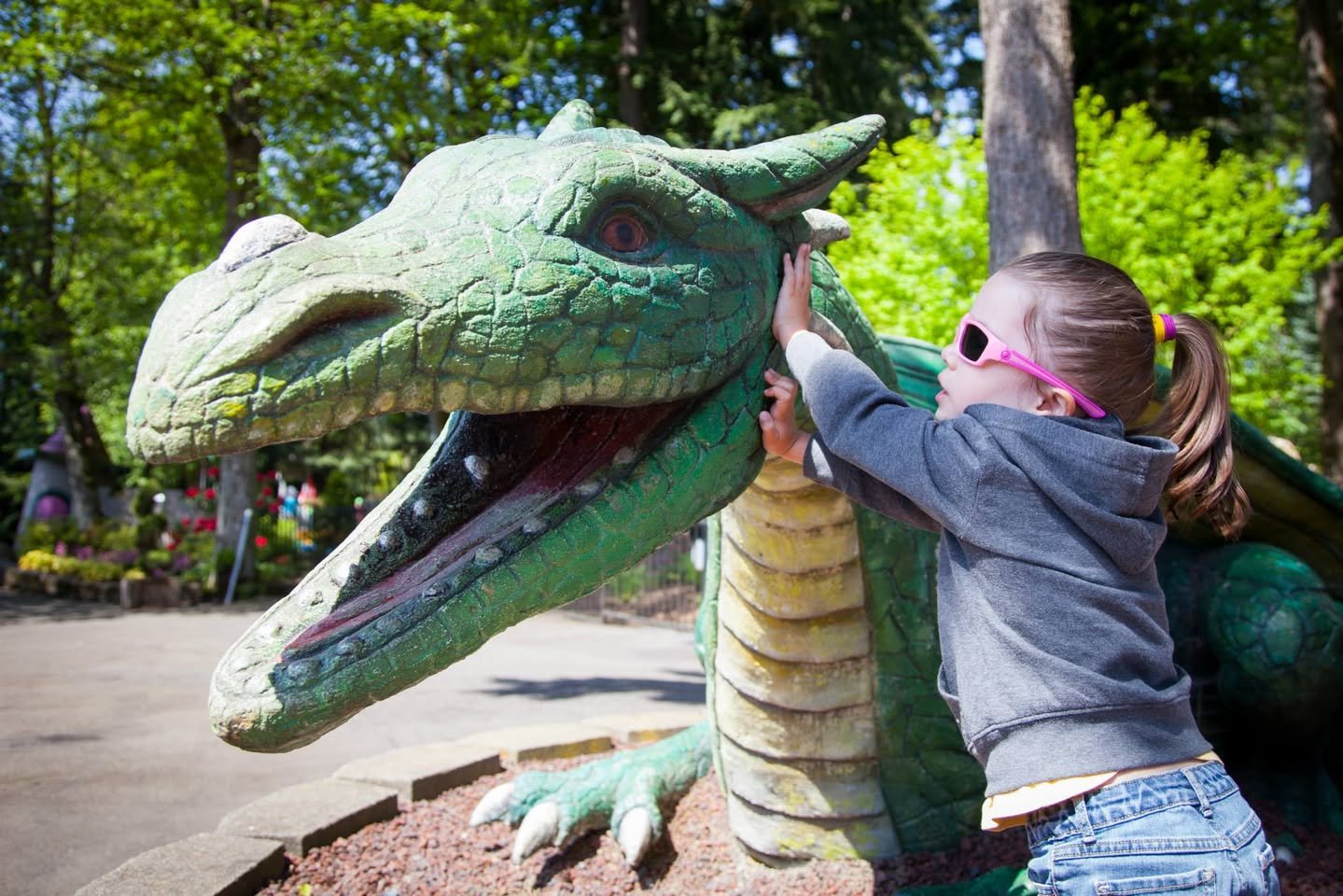 A child wears pink sunglasses and hugs a large green dragon statue in a sunny park setting.