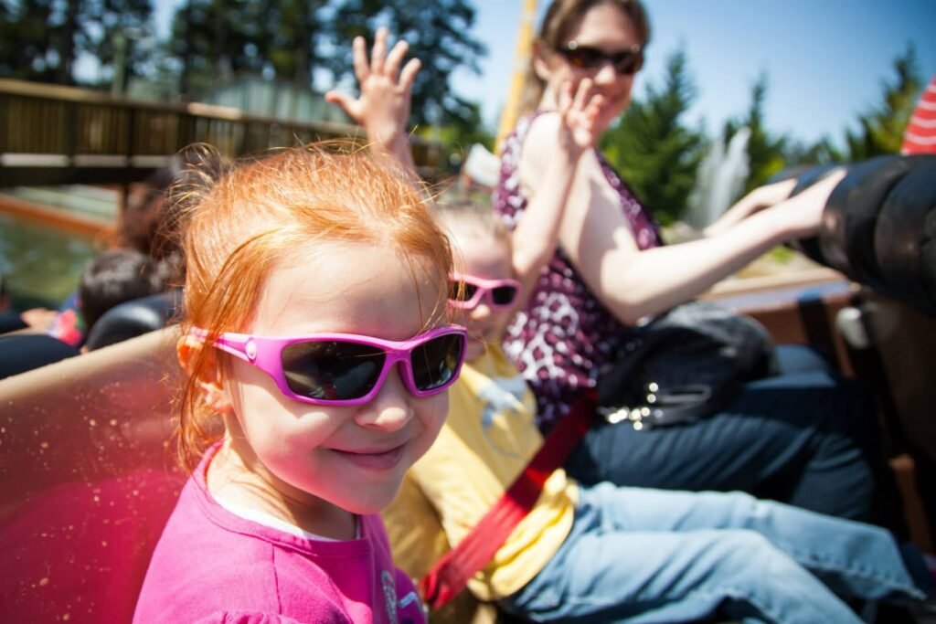 A smiling child wearing pink sunglasses sits on an outdoor ride with family members in the background.