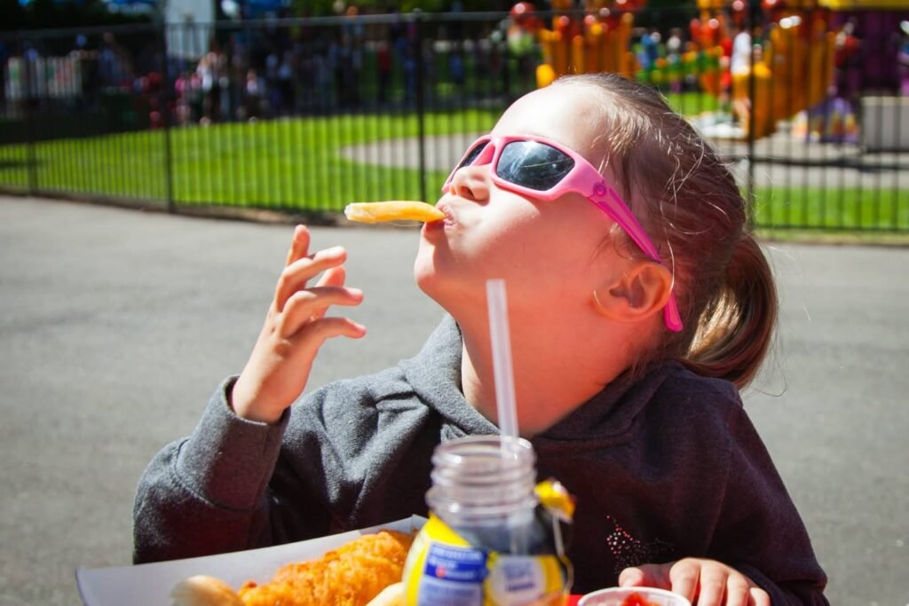 A young child wearing pink sunglasses eats a snack at an outdoor park on a sunny day.