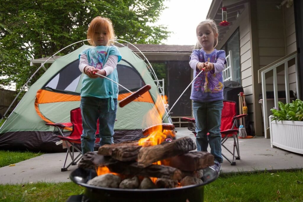 Two children roasting marshmallows over a fire pit while camping in a backyard tent.