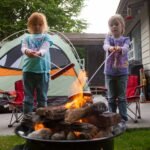 Two children roasting marshmallows over a fire pit while camping in a backyard tent.
