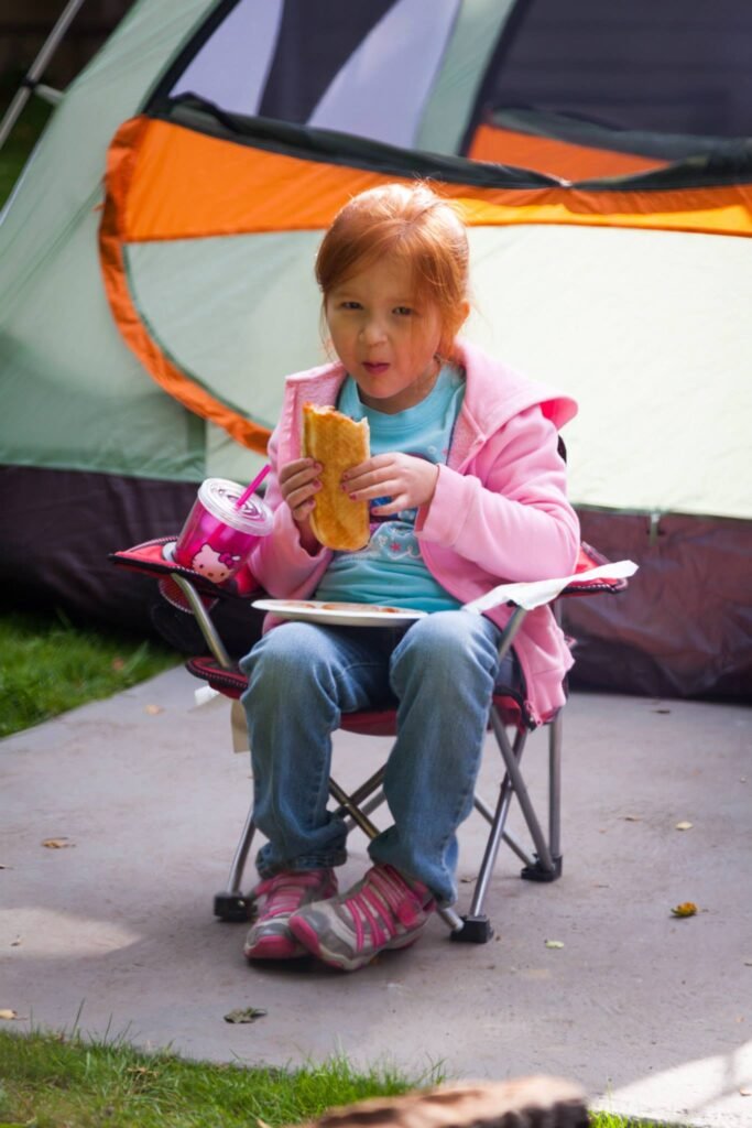 A young person in a pink jacket sitting in a folding chair eating in front of a tent.
