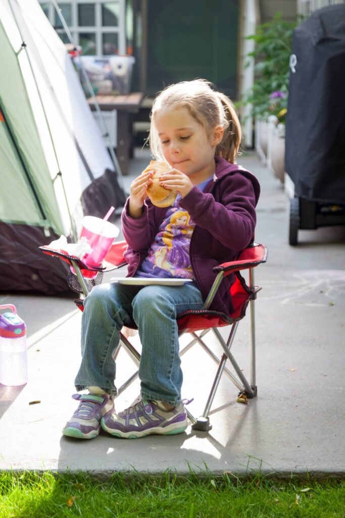 A child enjoying a snack while seated on a red folding chair outdoors. A green tent is visible in the background.
