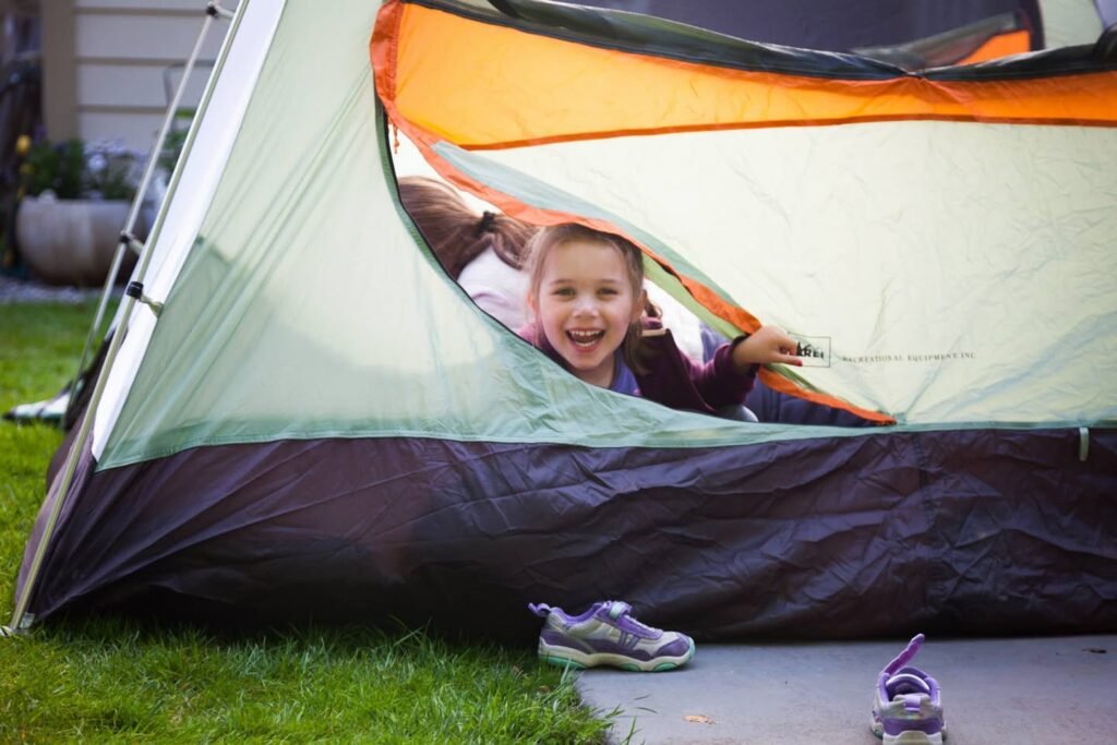 A child smiles from inside an orange and green camping tent set up on a lawn. A pair of sneakers rests near the tent entrance on a concrete path.