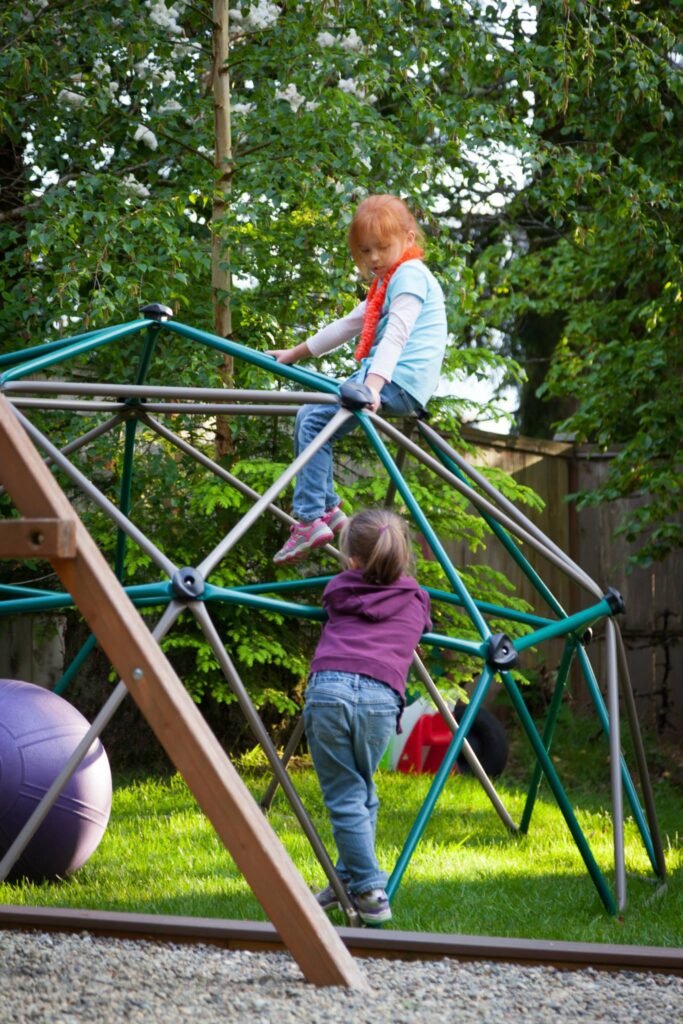 Two children climbing on a green metal and wood play structure in a grassy backyard.