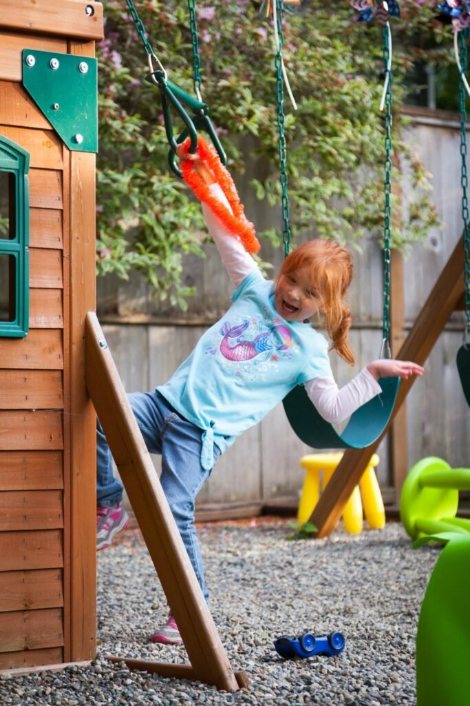 Children playing on a wooden swing set with a water bucket in a backyard garden.