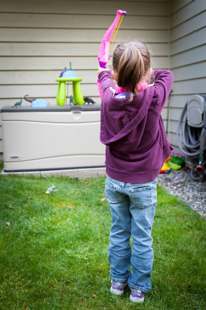 A child aims a pink toy bow toward a green bird feeder in a grassy backyard next to a hot tub.