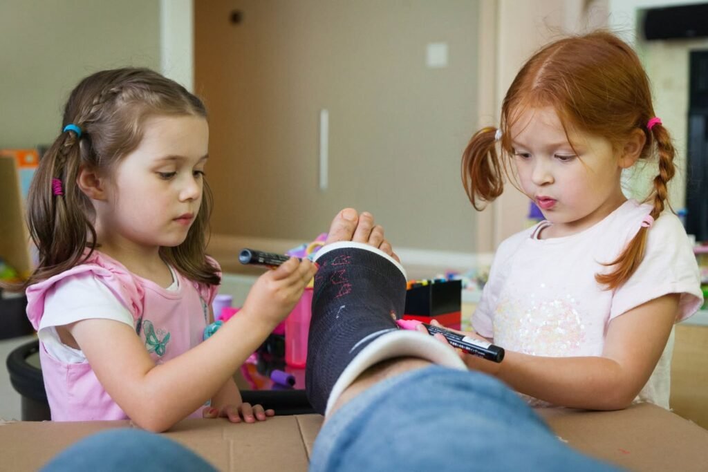 A photo of two young children using colored markers to decorate an adult's foot while at a kitchen table.