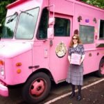 A woman in a grey skirt stands proudly in front of a large pink truck with decorations, holding a pink clipboard.