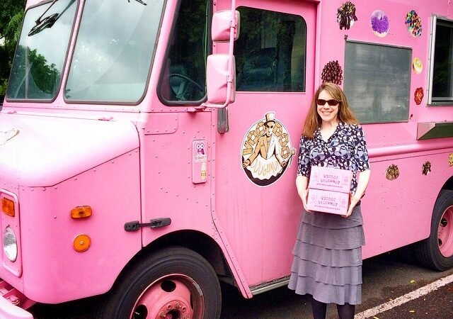 A woman in a grey skirt stands proudly in front of a large pink truck with decorations, holding a pink clipboard.
