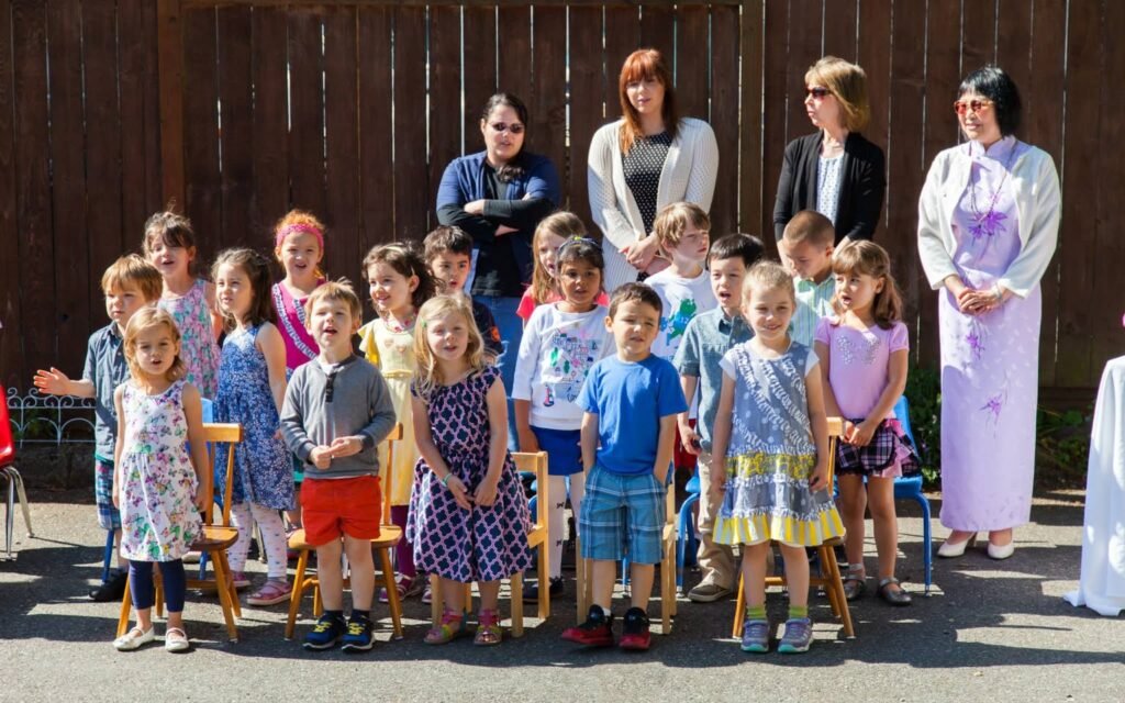 A group of children and adults are gathered outdoors for a formal photo session in front of a wooden fence.