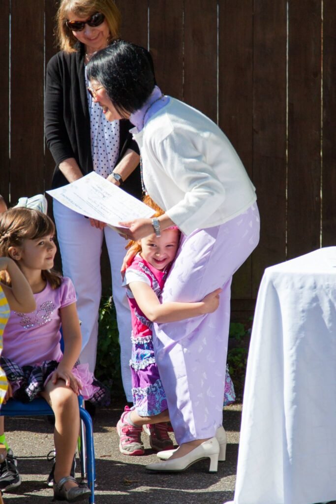 A woman in a white blazer holds a child joyfully, while others watch from a patio.